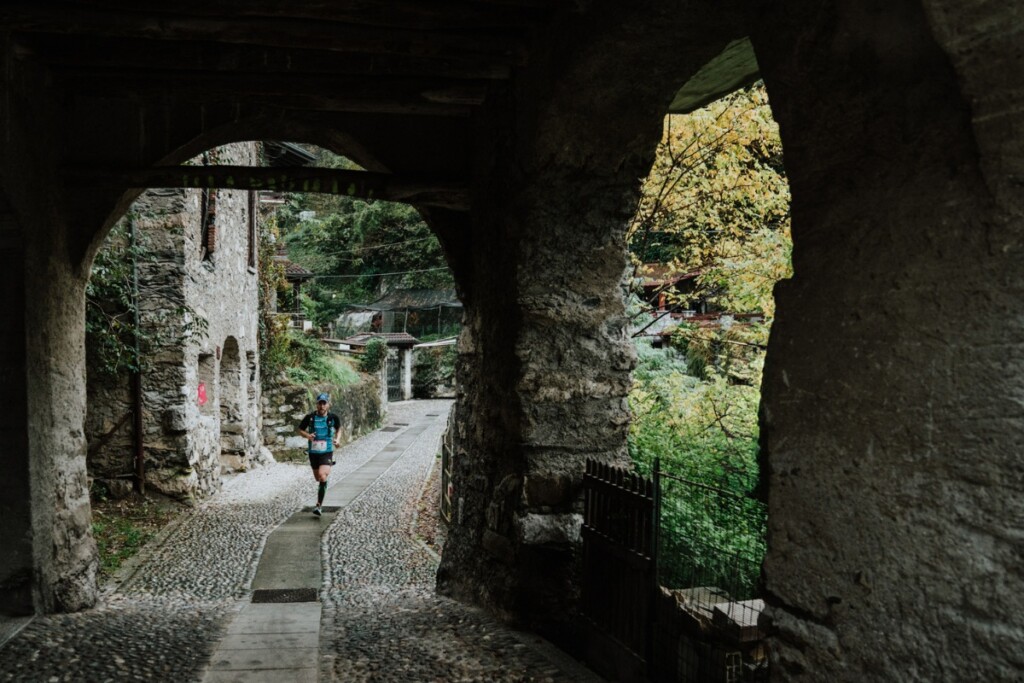 Trail runner attraversa un borgo storico in pietra a Losine durante la Valle dei Segni Wine Trail, immerso in un paesaggio autunnale della Valle Camonica.