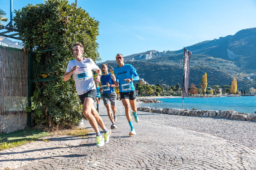 Runner in azione lungo il lago di Garda durante la Garda Trentino Half Marathon 2024, con sfondo di Riva del Garda e le montagne del Trentino.