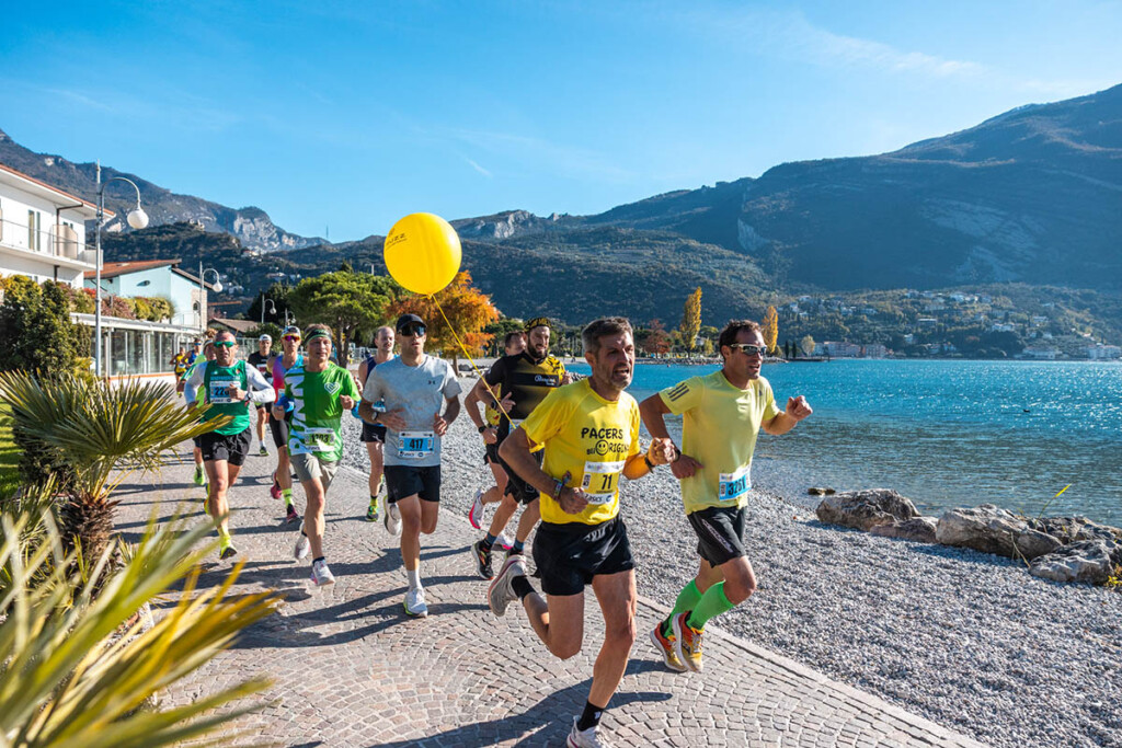 Gruppo di runner e pacer in corsa lungo il lago di Garda durante la Garda Trentino Half Marathon 2024, con sfondo di montagne e cielo limpido.