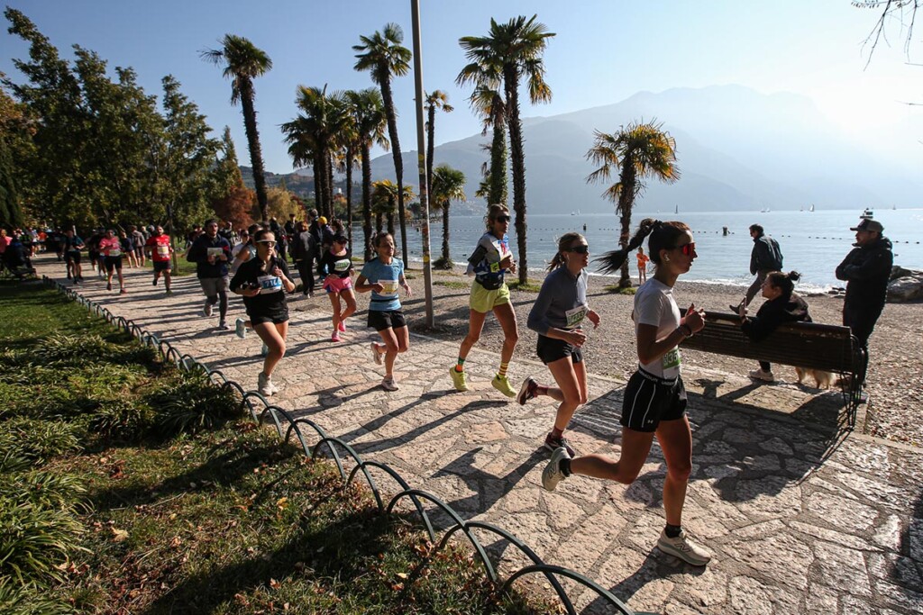 Gruppo di runner in azione sul lungolago di Riva del Garda durante la Garda Trentino Half Marathon 2025, con il lago e le montagne sullo sfondo.