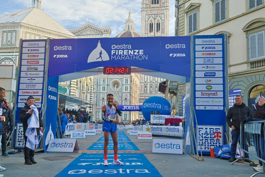 Badr Jaafari taglia il traguardo della Firenze Marathon 2025 in Piazza Duomo, sotto l’arco ufficiale Estra con il cronometro visibile e la Cattedrale di Santa Maria del Fiore sullo sfondo.