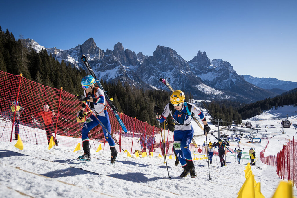Atleti Rocco Baldini e Giovanni Rossi impegnati nella salita della sprint di sci alpinismo a Madonna di Campiglio, con le cime dolomitiche sullo sfondo e rete di protezione lungo il tracciato.